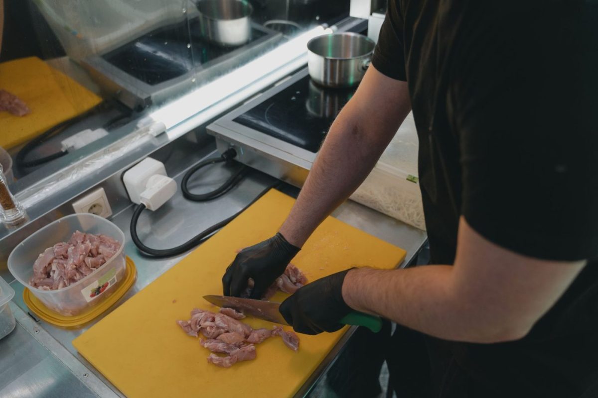 A cook in a commercial kitchen feeds meat into a large stainless steel industrial meat grinder.