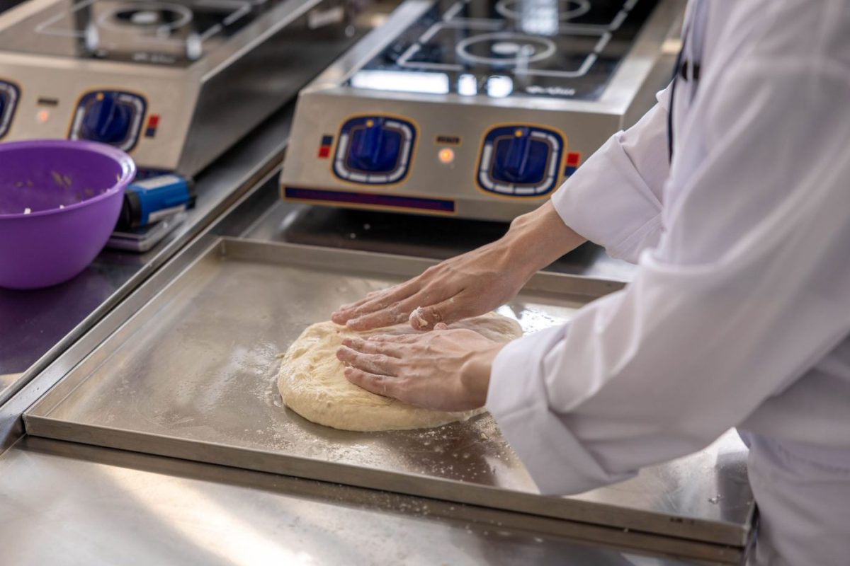 A baker in a commercial kitchen checks the temperature of dough in a large spiral mixer to prevent overheating.